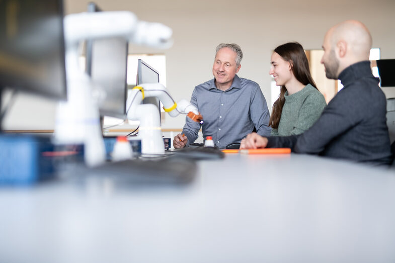 <br>Photo of a teacher and two students. They are sitting in front of a PC and focus their eyes on the monitor. On the left are other PCs and a swivel-arm robot. One student is taking notes on the tablet.<br>