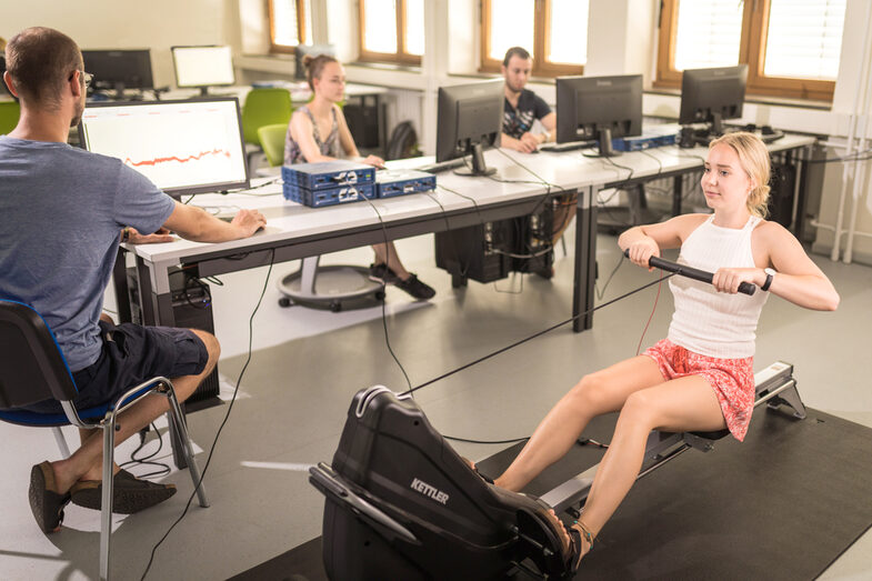 Photo of a young woman in action on a rowing machine. To her left and in the background you can see three people sitting at a PC and taking measurements.