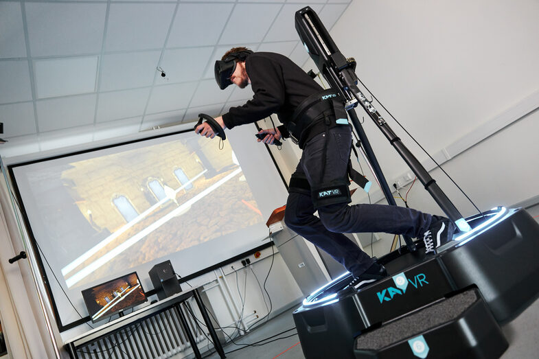 Photo of a man wearing VR goggles and holding controllers in his hands. He is standing on a device that allows him to move on the spot. Next to him, a projector screen shows what he is currently seeing.